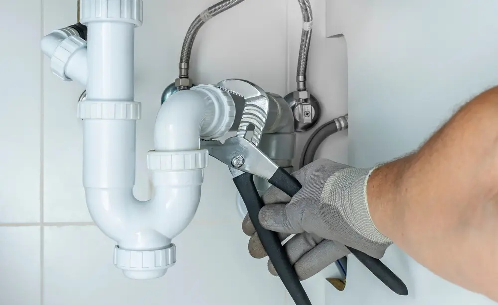 Gloved hand adjusting a white sink drain trap with pliers