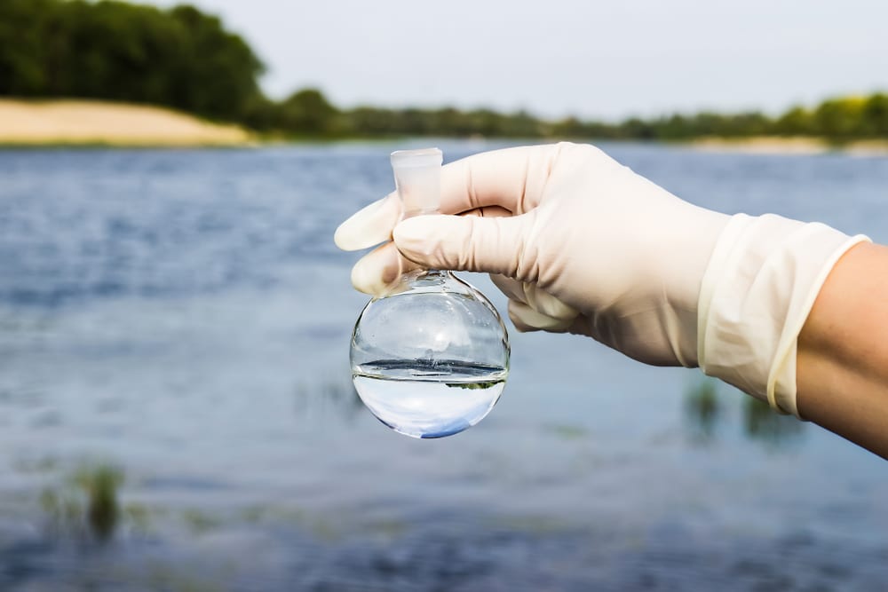 Gloved hand holding a water sample flask above a lake