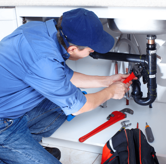 Plumber in a blue cap repairing pipes under a sink with a red wrench