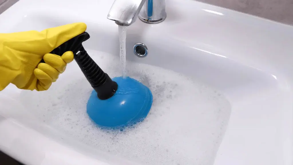 Gloved hand using a plunger on a bathroom sink drain