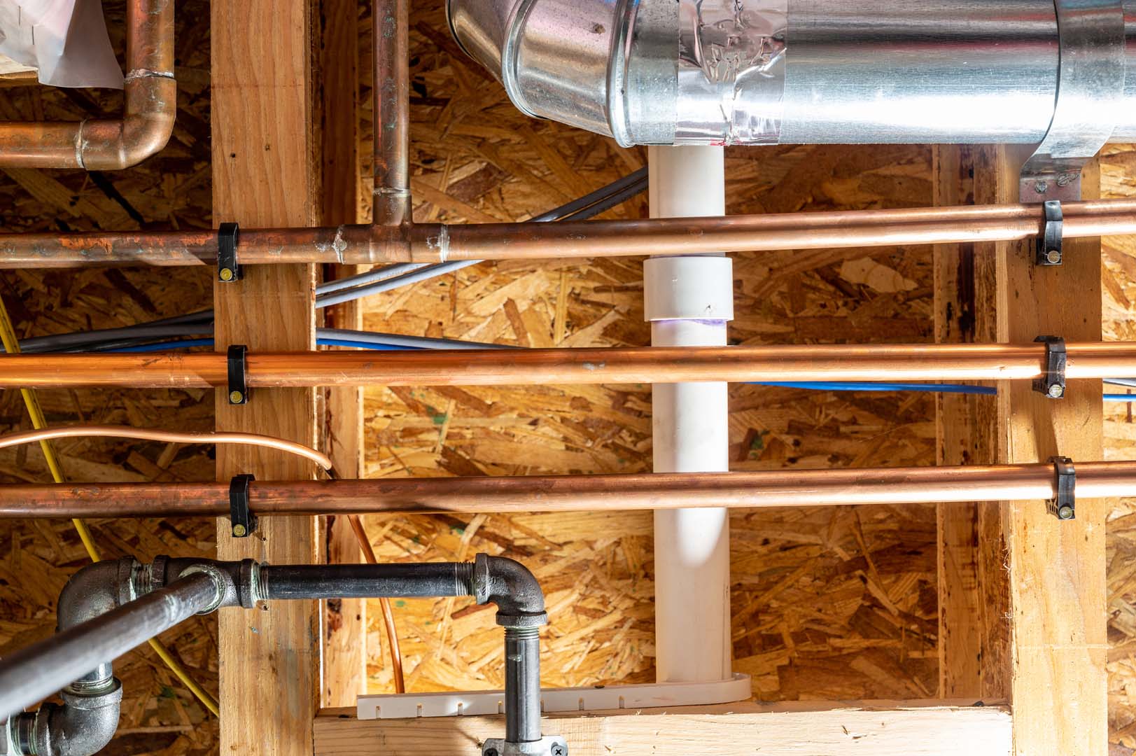 Copper water lines, a black gas pipe and a PVC drain running through exposed floor joists