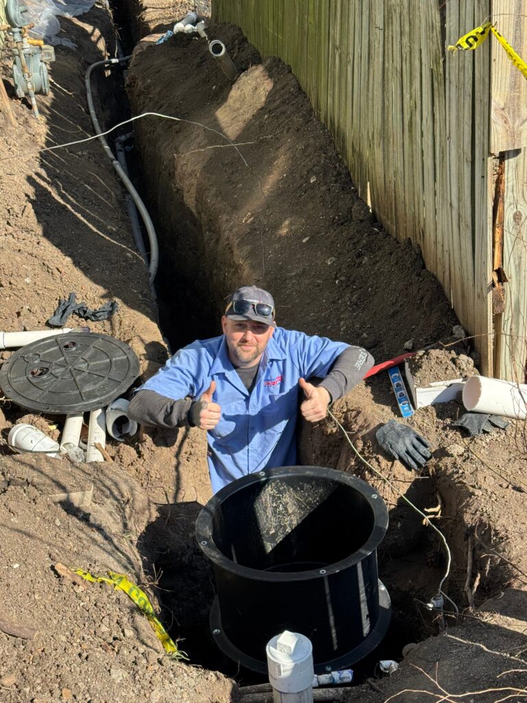 Smiling plumber giving thumbs up in a trench beside a new black sewer basin